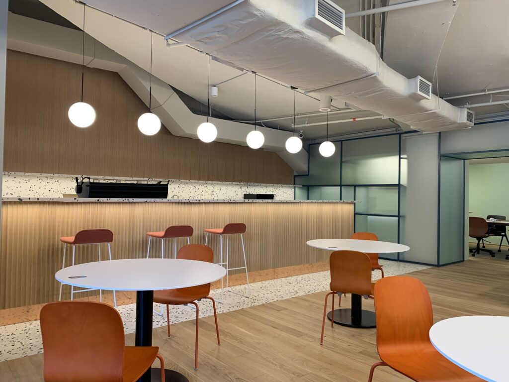 A contemporary modern interior fit-out for a coworking space featuring orange wood-tone chairs and feature lights overhung on the kitchen area