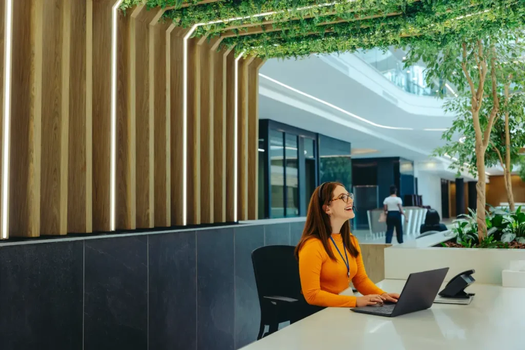 A friendly receptionist with a bright smile sits at a desk using a laptop in a modern office lobby. The setting features natural elements and contemporary design, creating a welcoming atmosphere.